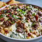 Golden, bubbly Philly Cheesesteak Dip in a baking dish, ready to serve with toasted baguette slices. 