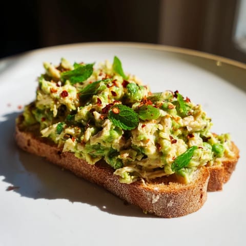 A close-up view of a warm Smashed Pea & Chicken Toast garnished with fresh mint, lemon zest, and a drizzle of olive oil.  