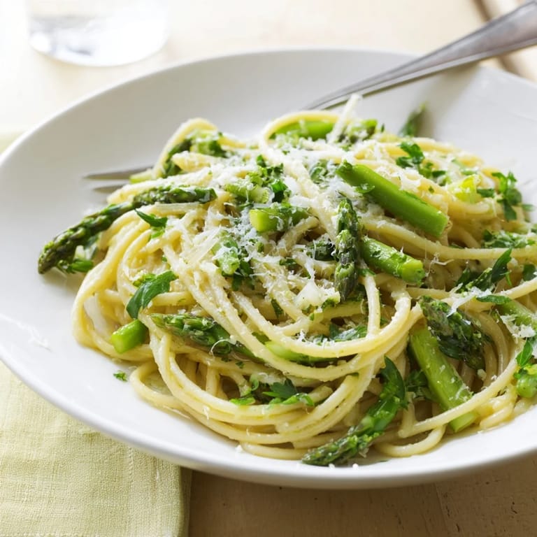 A close-up shows tender asparagus pieces and mashed roasted garlic cloves mixed with spaghetti, finished with lemon zest and a drizzle of olive oil.